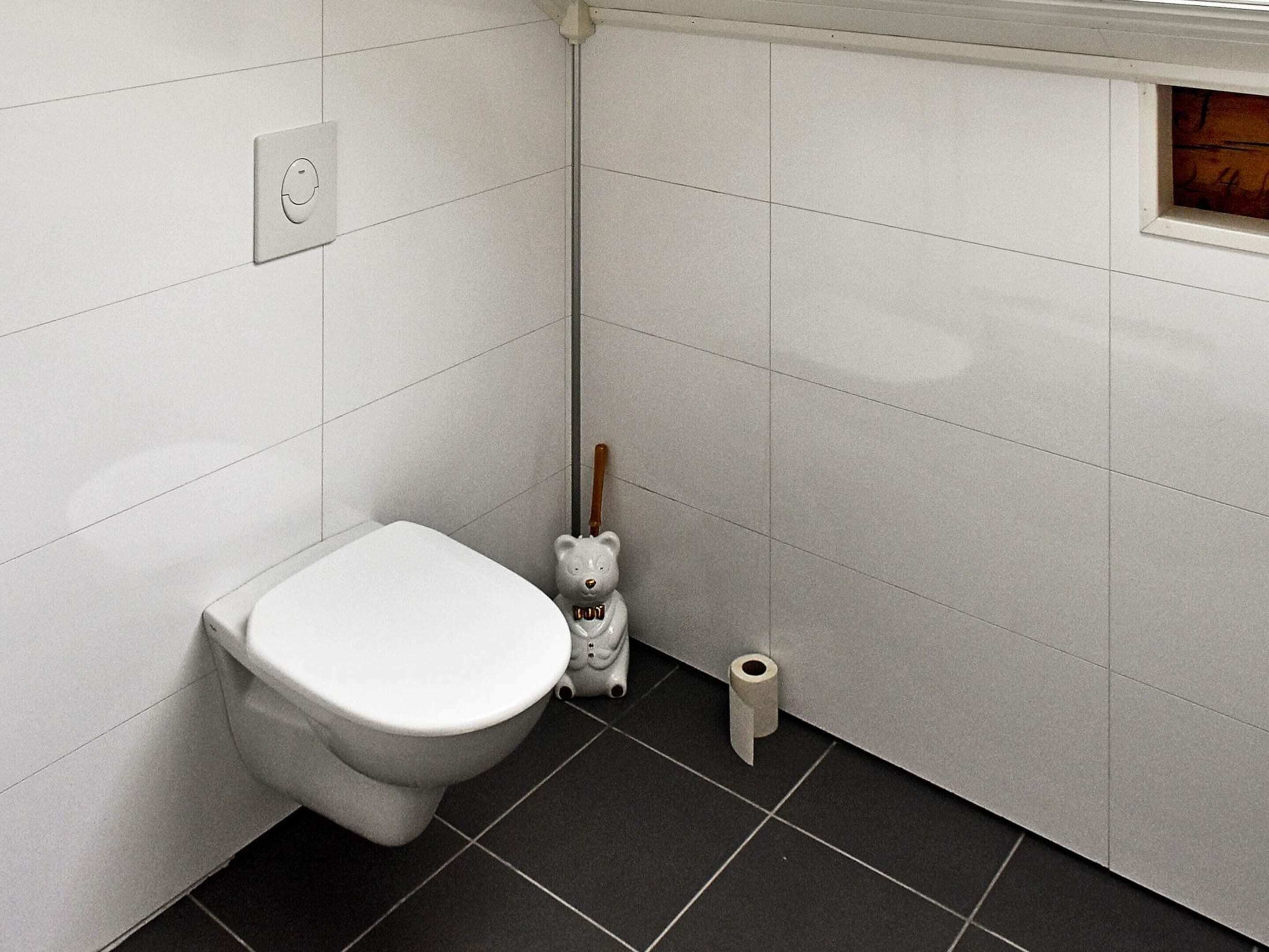 A toilet with a bear-shaped bidet attachment, a roll of toilet paper, and a toilet brush.
