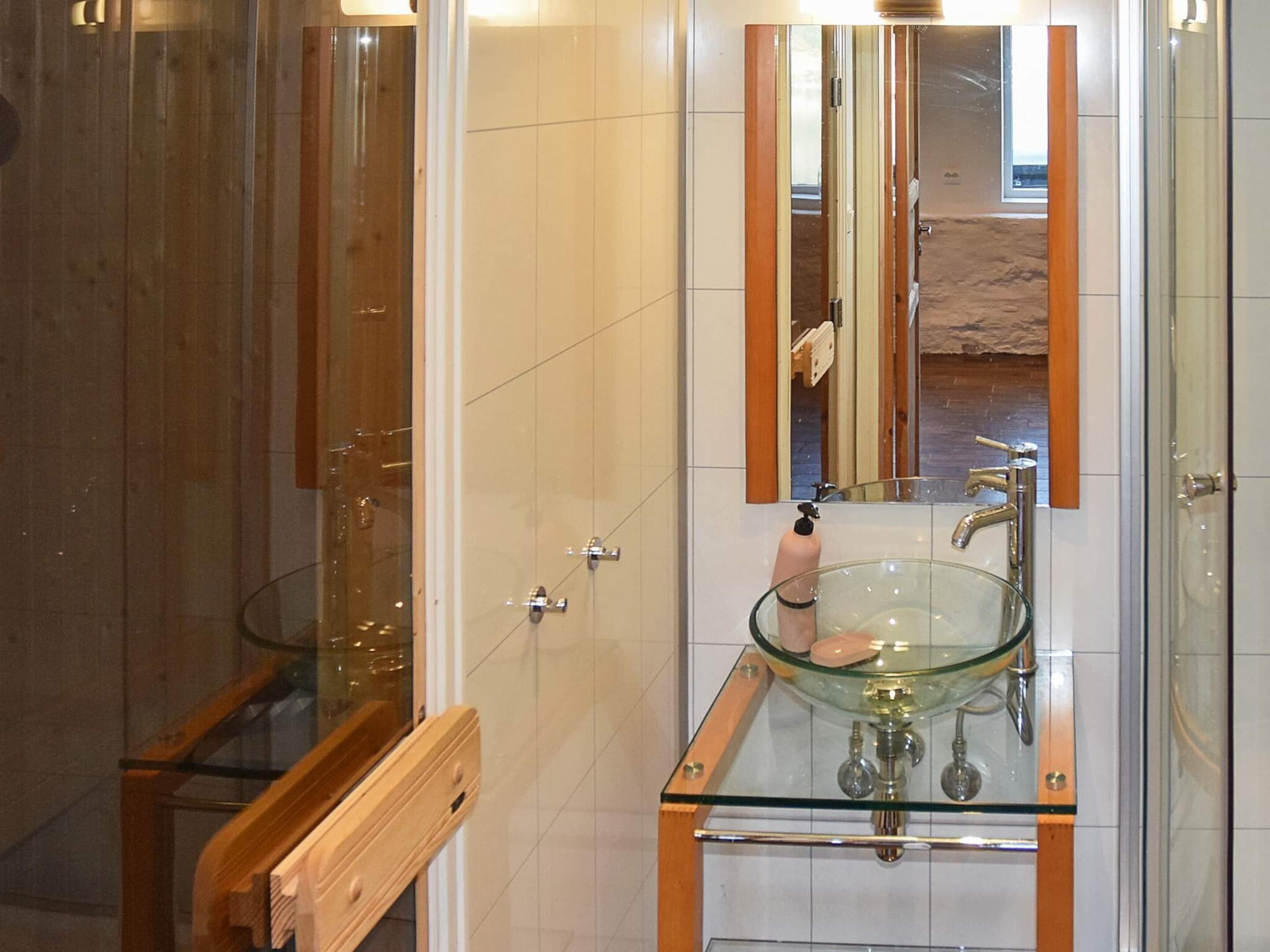 A bathroom with a glass sink, a wooden vanity, a mirror, and a shower area.