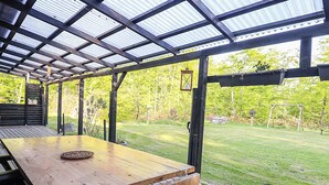 An outdoor wooden deck with a table and chairs, surrounded by a metal roof structure and a view of a grassy yard with trees. - 6 Person Holiday Home in Hadsund-by Traum (Hadsund)