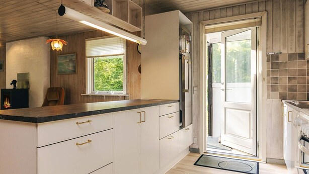 A kitchen with white cabinets, a black countertop, and a wooden ceiling. There is a window with blinds, a door, and a small shelf with decorative items. - 6 Person Holiday Home in Hadsund-by Traum (Hadsund)
