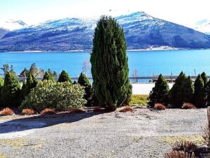 Ein Kiesgrundstück mit einer hohen Nadelbaum, einem Gewässer und verschneiten Bergen im Hintergrund.
