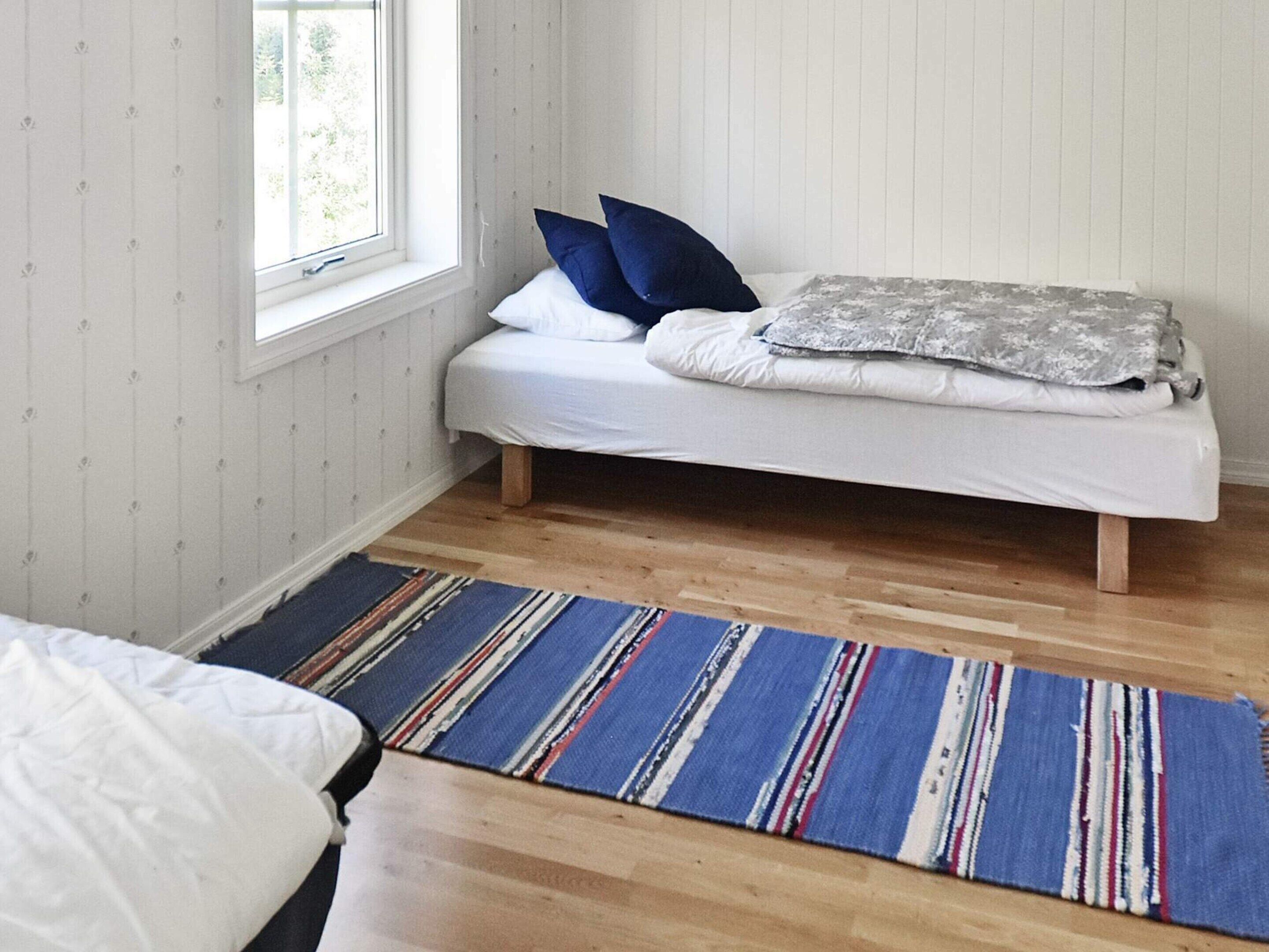 A single bed with a white mattress and blue pillows, a gray blanket, and a striped rug on a wooden floor.