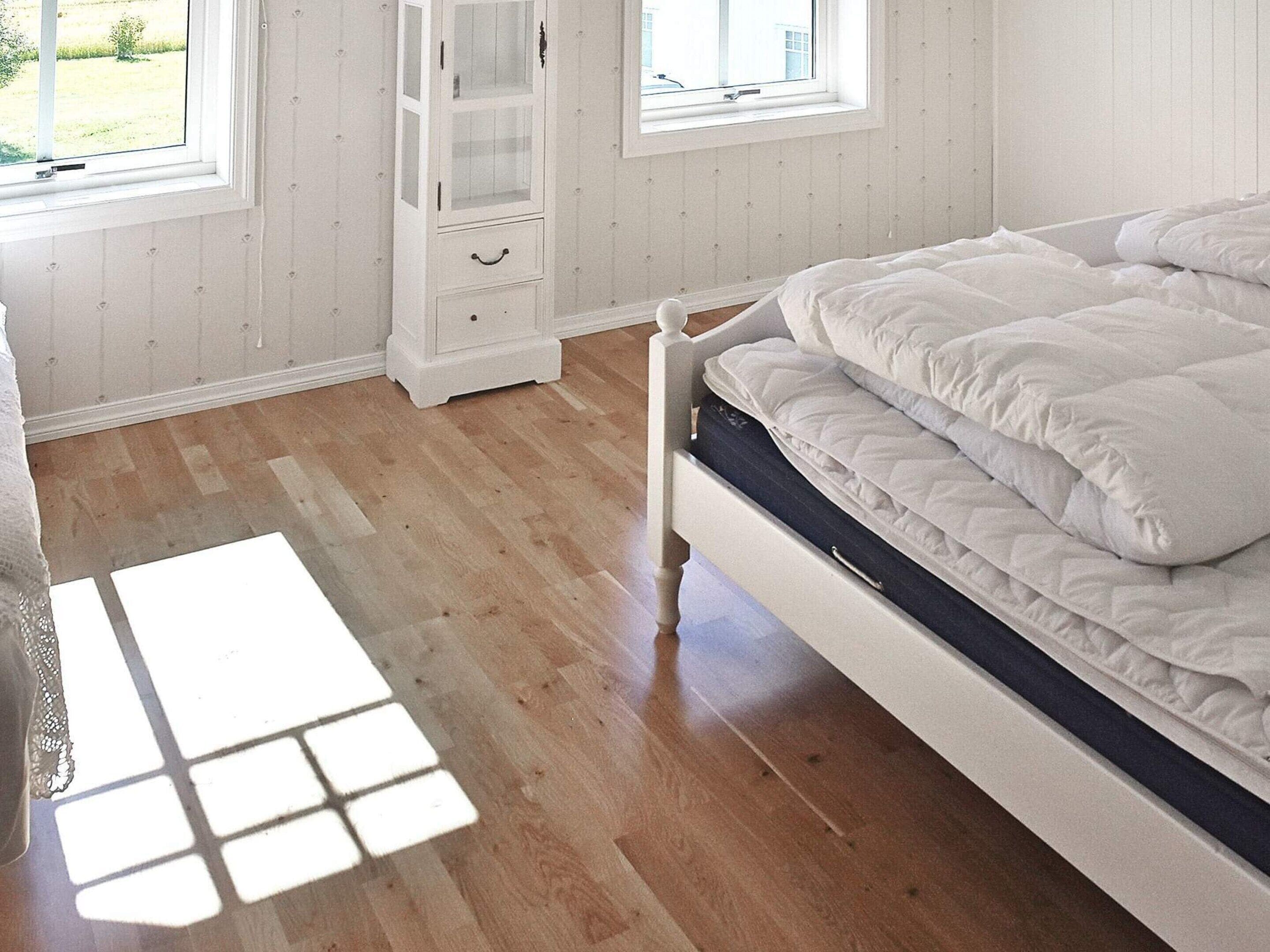 Un dormitorio con piso de madera, una cama con ropa blanca, un armario blanco y un ventanal con vista a la vegetación.