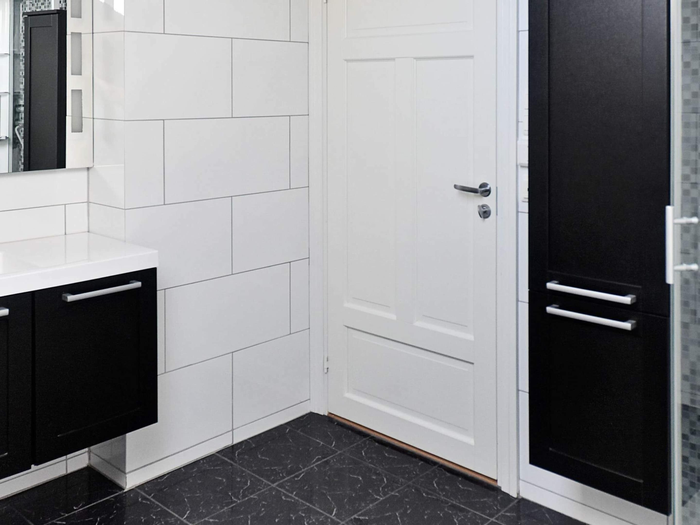A bathroom with white tiled walls, a black tiled floor, a white door with a silver handle, and black cabinets with silver handles.