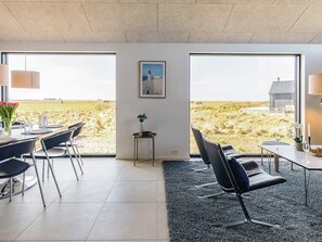 A modern interior with a dining area, two large windows offering a view of a grassy field, and a small table with a vase of flowers.