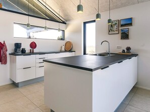 A modern kitchen with a central island, white cabinetry, a built-in oven, and a window providing natural light. - 5 Star Holiday Home in Lokken (Lokken)