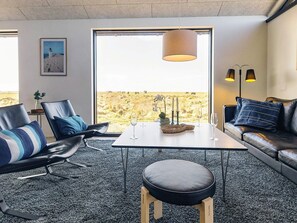 A modern living room with a view of a natural landscape, a dining table with glasses, and a sofa with cushions.