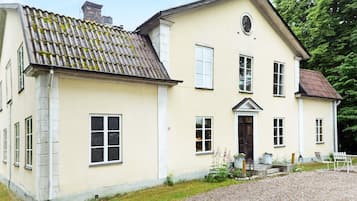A two-story building with a symmetrical facade, a central entrance with a door and windows on either side, and a gravel driveway leading to a seating area with outdoor furniture.