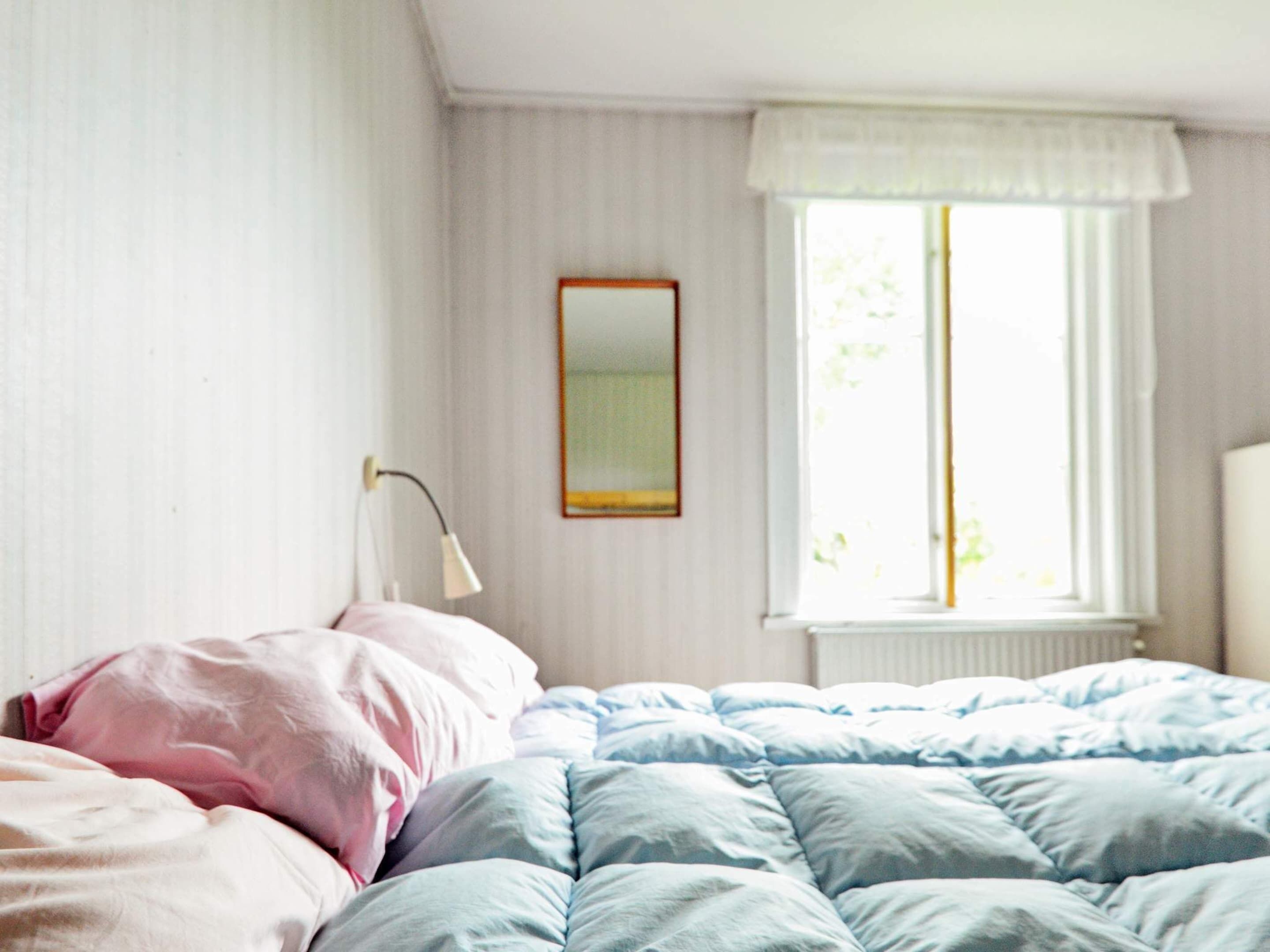 A bedroom with a bed, a bedside lamp, a window with curtains, and a mirror on the wall.