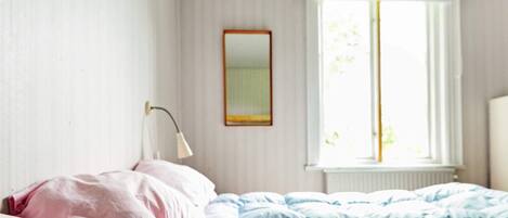 A bedroom with a bed, a bedside lamp, a window with curtains, and a mirror on the wall.