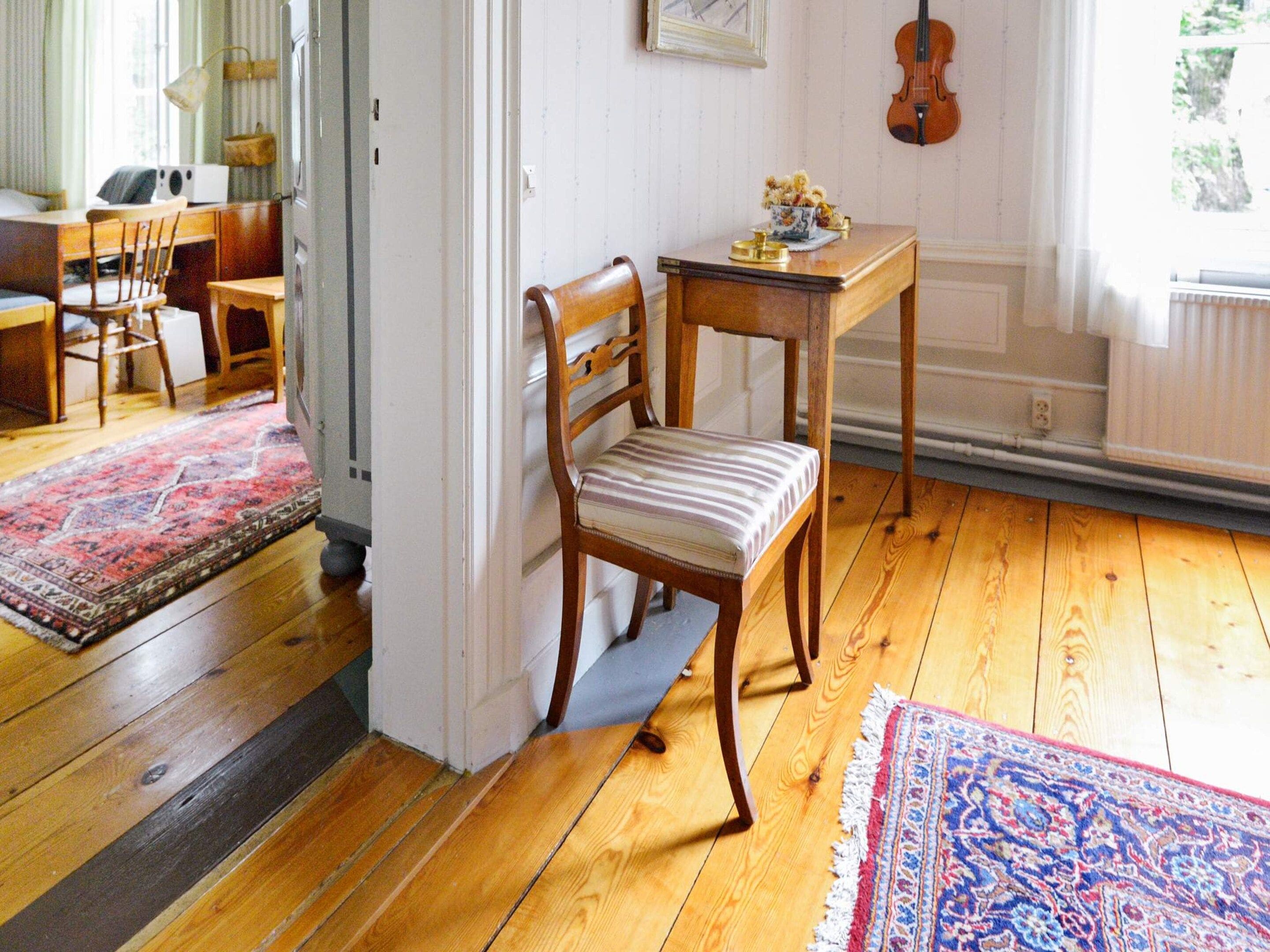 Una habitación con piso de madera, una silla a rayas, una pequeña mesa con un florero y un violín en la pared.