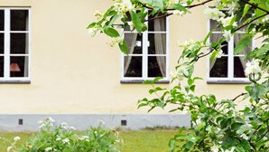 Ein Gebäude mit hellgelber Fassade, drei Fenstern mit weißen Rahmen und einem Garten mit weißen Blütenpflanzen.