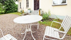 Ein weißer Sitzbereich im Freien mit einem runden Tisch und zwei Stühlen, umgeben von Kies, vor einem gelben Gebäude mit einer dunklen Tür und Fenstern.