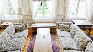 Ein geräumiges Wohnzimmer mit einer Chandelieber, zwei Sofas, einem Couchtisch und großen Fenstern mit Vorhängen.