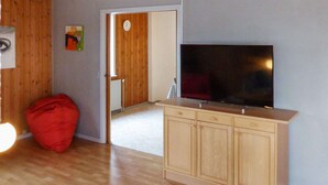 Una habitación con suelo de madera, un televisor sobre un mueble, un sillón de peluche rojo y una puerta que da a otra habitación.