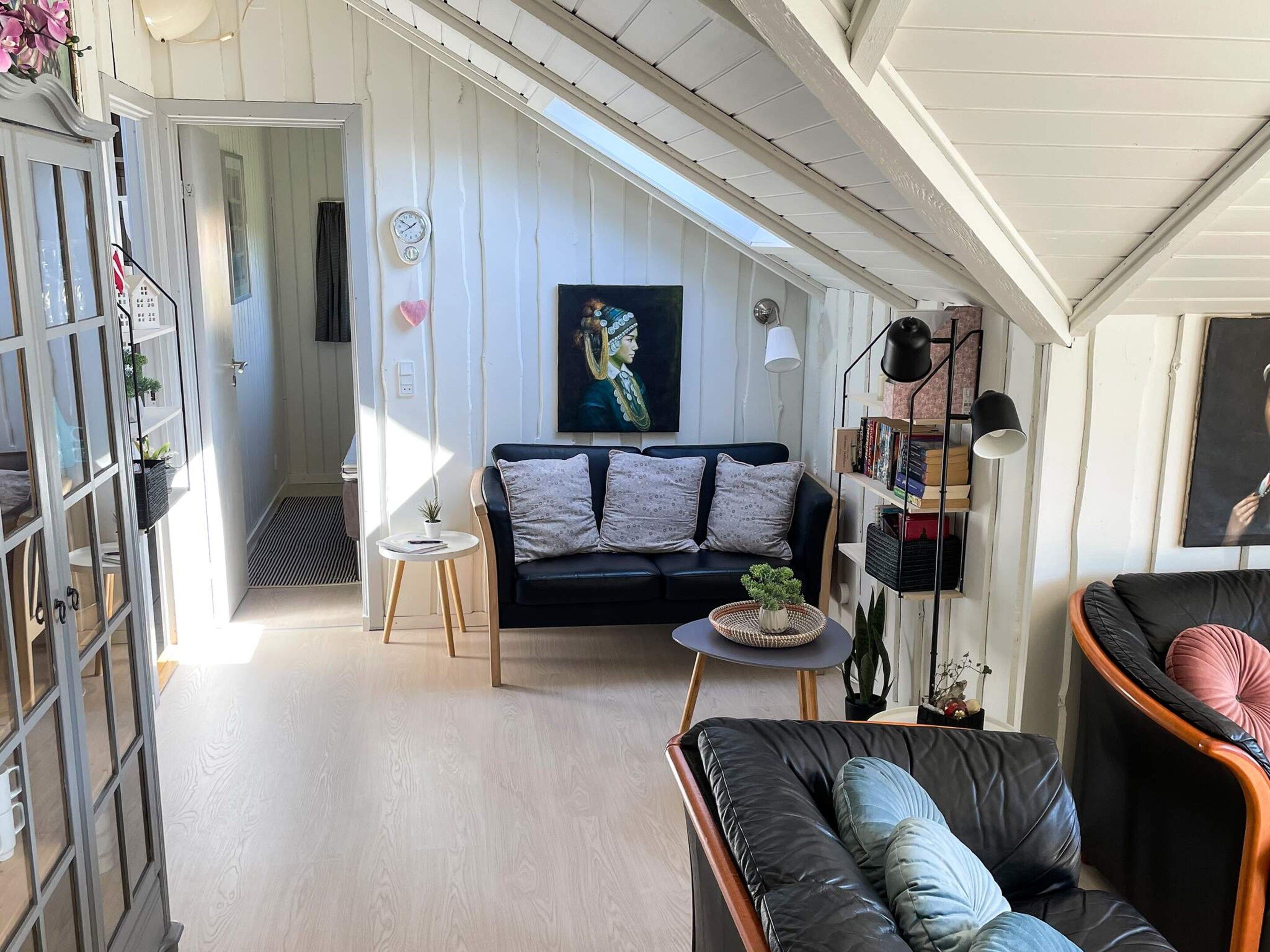 A cozy living room with a black sofa, a black leather chair, and a small round table. There is a bookshelf with books and a painting on the wall.