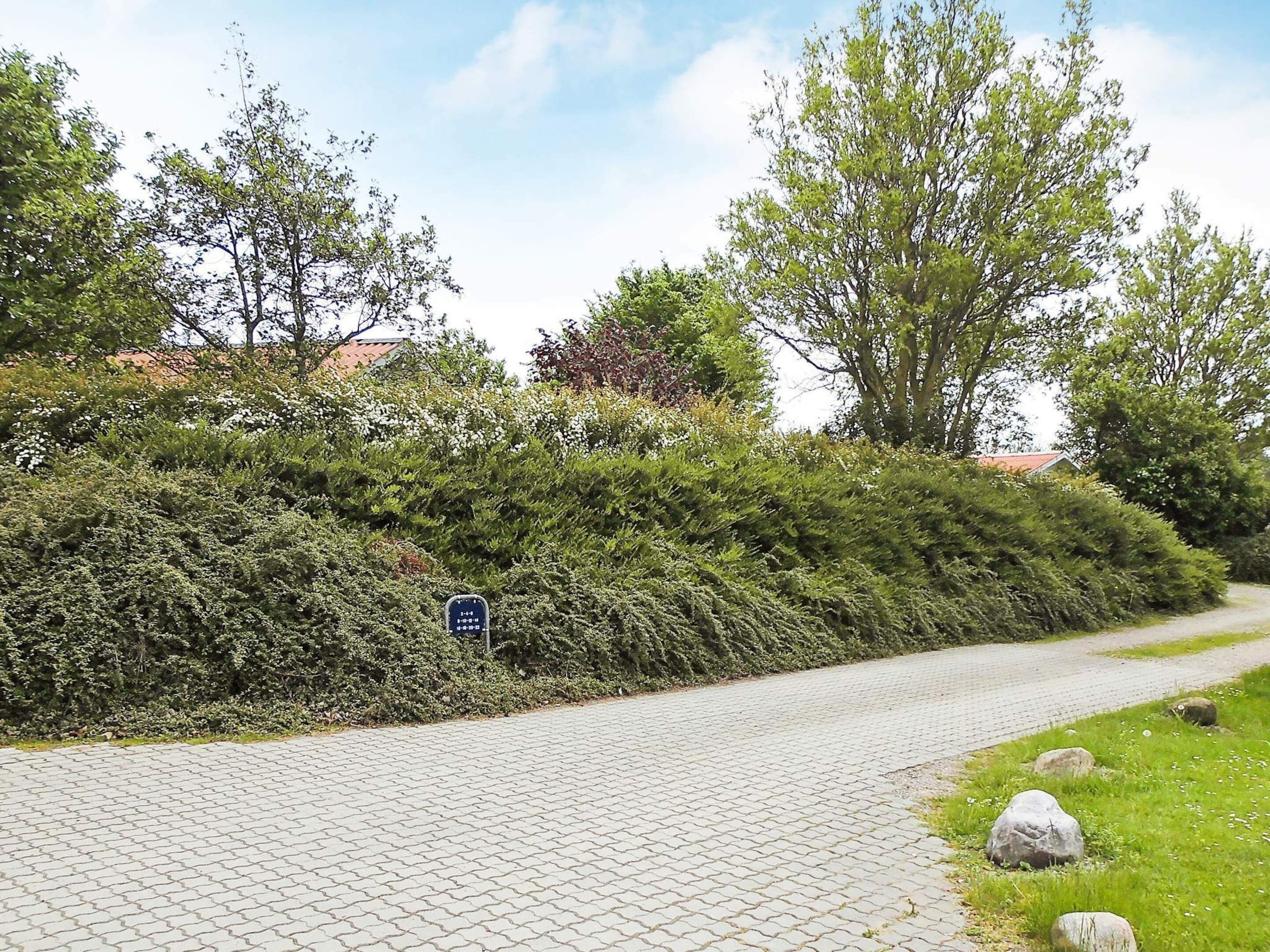 A paved pathway bordered by dense shrubbery and trees, leading to a building with a red roof.
