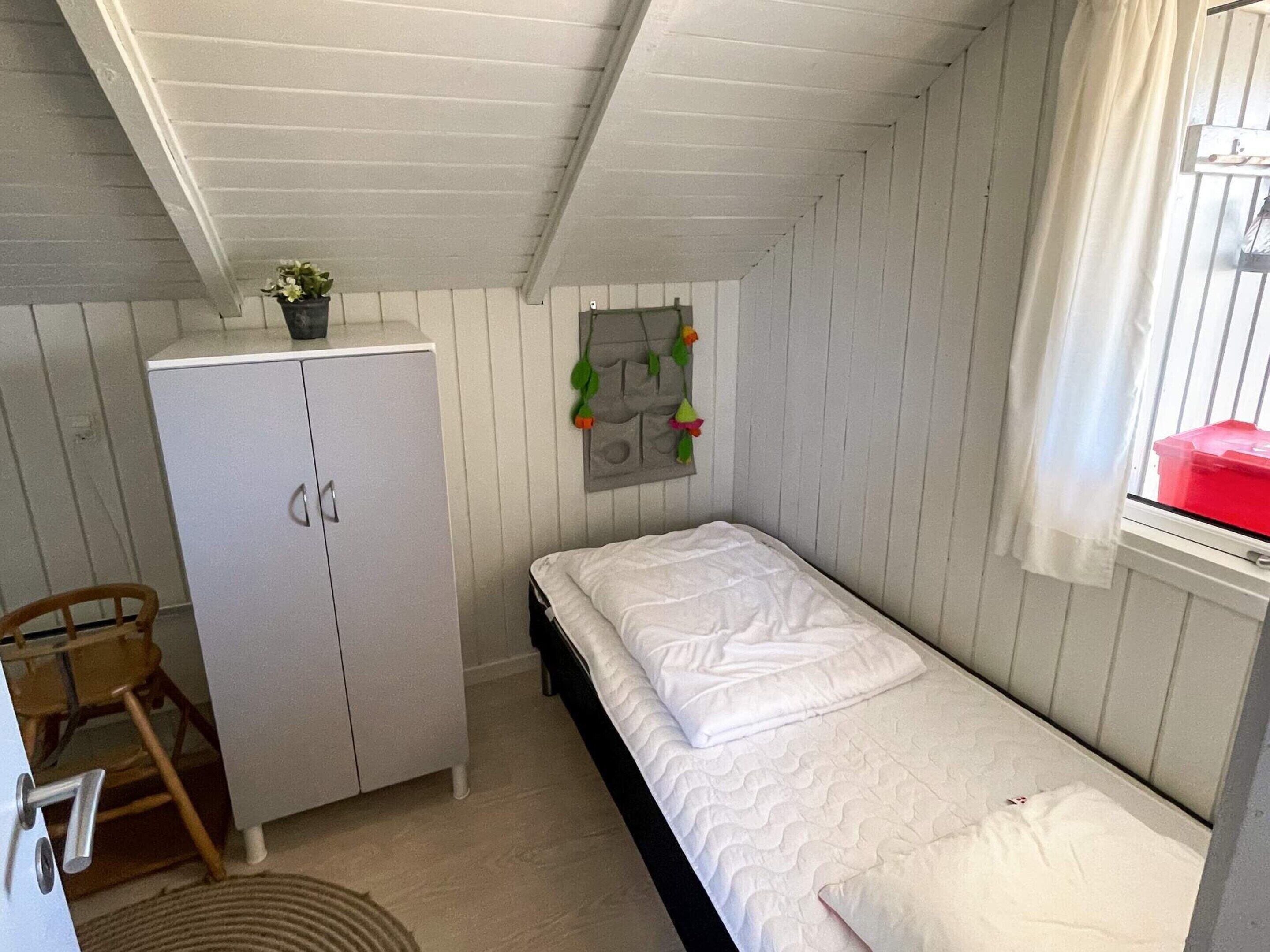 A small, single-bed room with a wooden wardrobe, a small table, and a window with a red container.