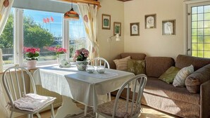 Ein gemütliches Zimmer mit Esstisch, Stühlen und einer Couch. An den Wänden befinden sich Kübelpflanzen und gerahmte Bilder.