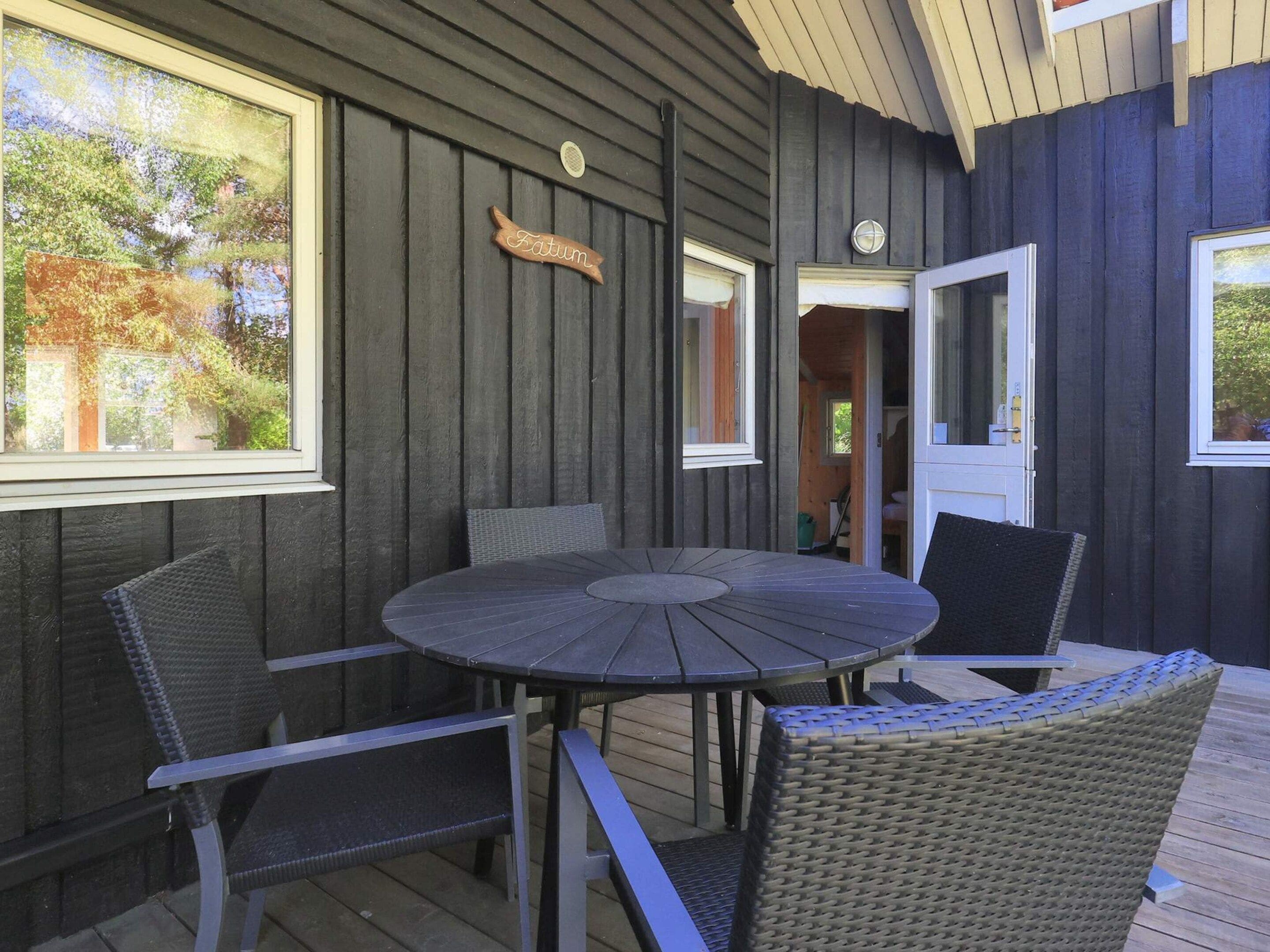 Una terraza de madera con una mesa redonda y cuatro sillas, una ventana con vista a árboles y una puerta que da a un espacio interior.