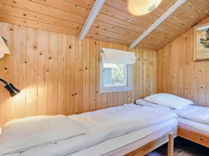 A wooden cabin room with two beds, a window with a white curtain, and a painting on the wall.