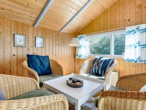 A cozy room with wooden walls, wicker furniture, a white table with a decorative bowl, and a window with curtains.