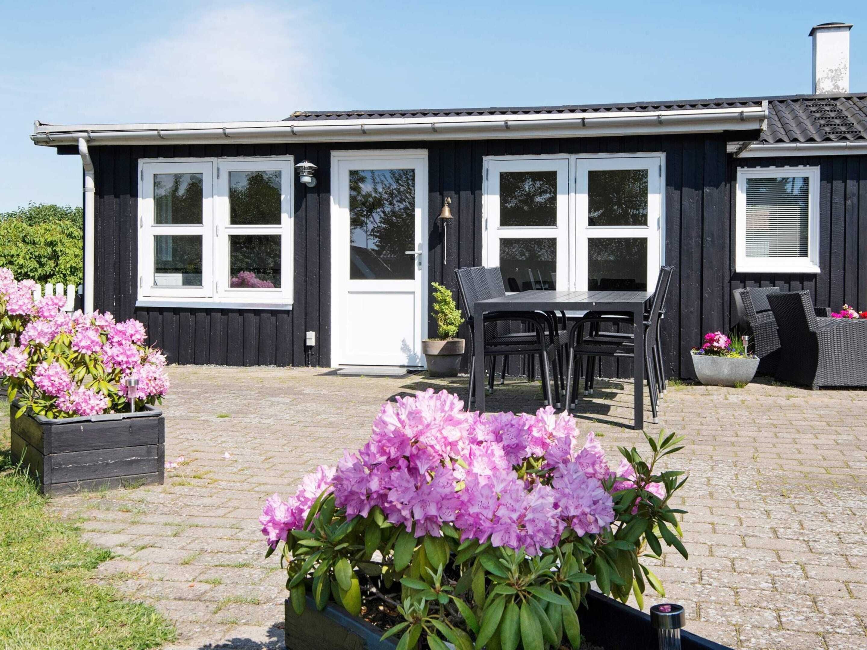 A black building with white-framed windows, a door, and a chimney. There is a patio with a table and chairs, and potted plants with pink flowers.