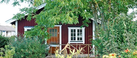 A red wooden house with a white door and windows, surrounded by lush greenery and fruit-bearing plants.