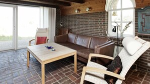 Ett mysigt rum med ett träpanelat tak, en tegelvägg, en brun lädersoffa, en vit fåtölj och ett träbord.