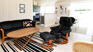 Ein modernes Wohnzimmer mit einem schwarzen Ledersofa, einem Holztisch und einem schwarzen Ledersessel. Im Hintergrund ist eine Küche mit weißen Schränken und einem Fenster zu sehen.