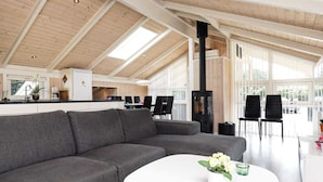 A modern interior with a grey sofa, a white coffee table with flowers, and a kitchen area with a stove. - 9 Person Holiday Home in Jerup (Jerup)