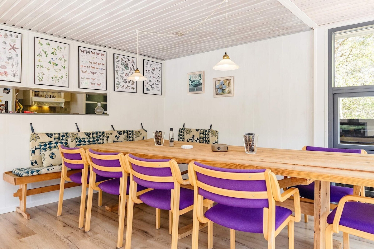 A dining area with a wooden table and purple chairs, white walls with framed botanical prints, and a window with a view of trees.