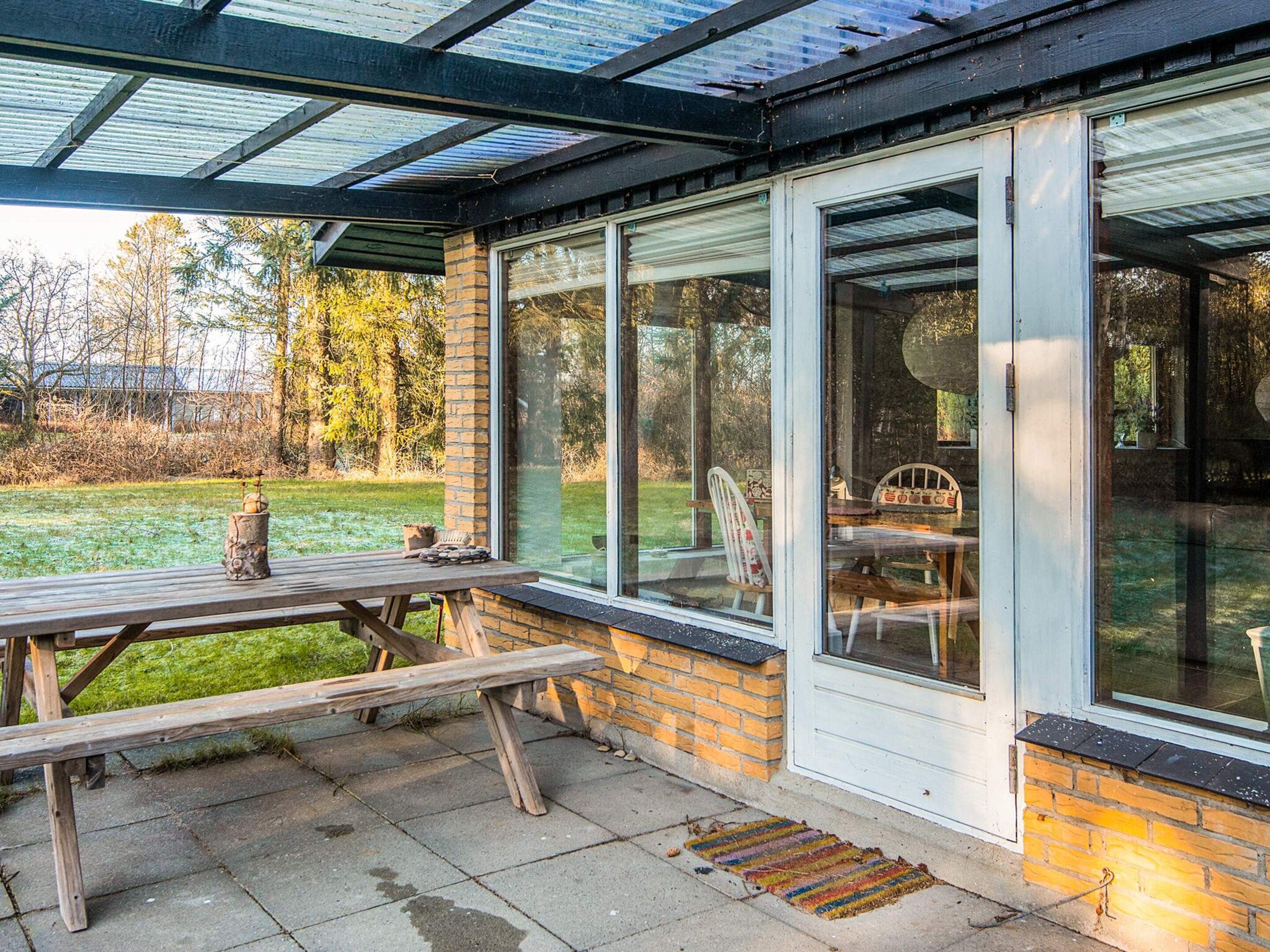 A covered patio with a wooden table and chairs, large windows, and a brick wall.