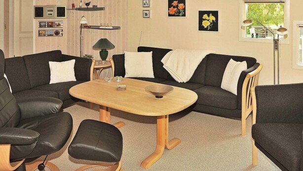 A living room with a sofa set, a coffee table, and a lamp. There are framed pictures on the wall and a window with a view of greenery.