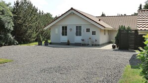 A small, single-story building with a white exterior and a tiled roof. There is a gravel driveway leading to the entrance, and two chairs are placed outside the building.