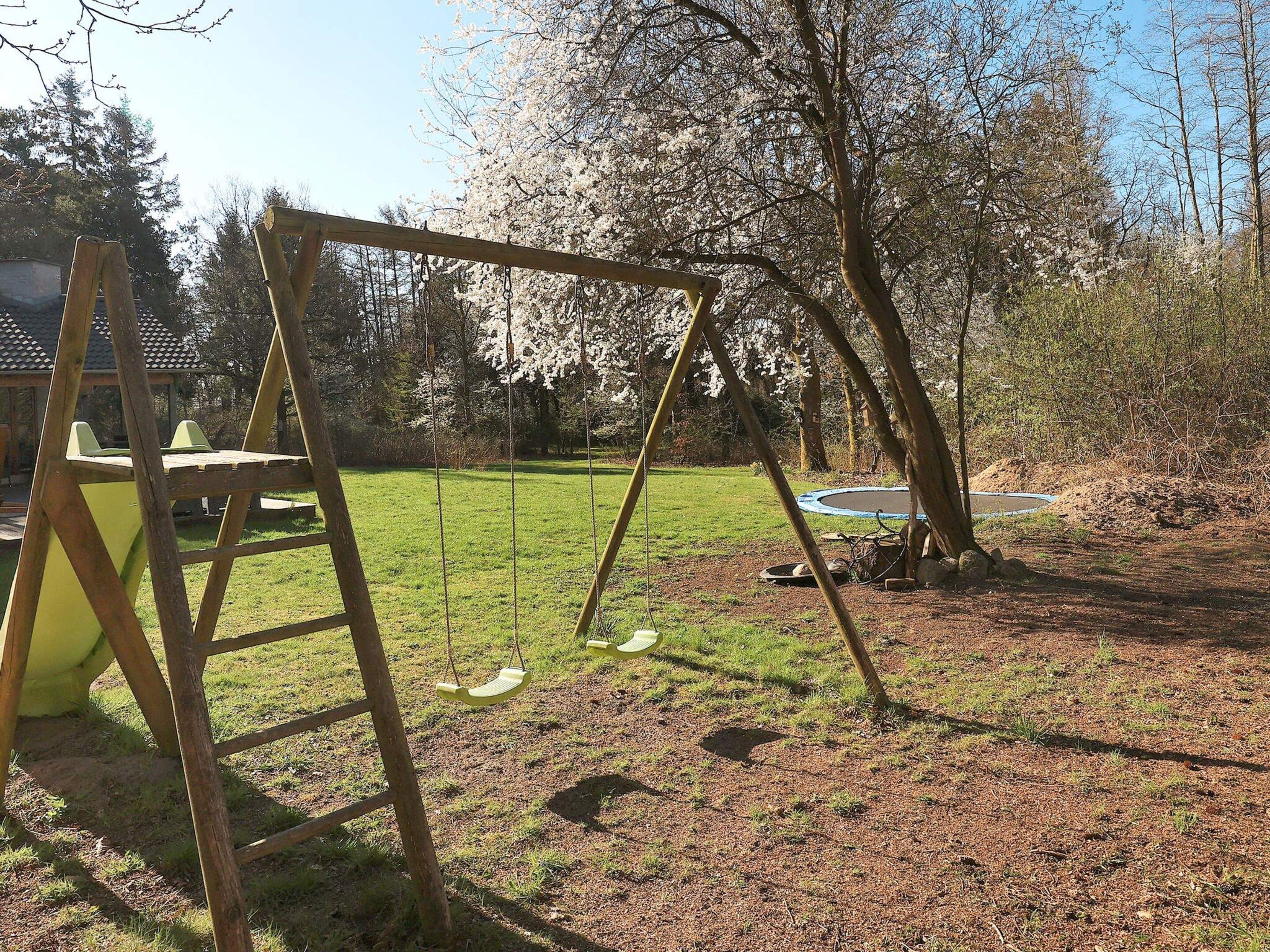 Un ensemble de jeux d’extérieur en bois, avec une balançoire, installé dans une cour gazonnée, avec un trampoline et des arbres en arrière-plan.