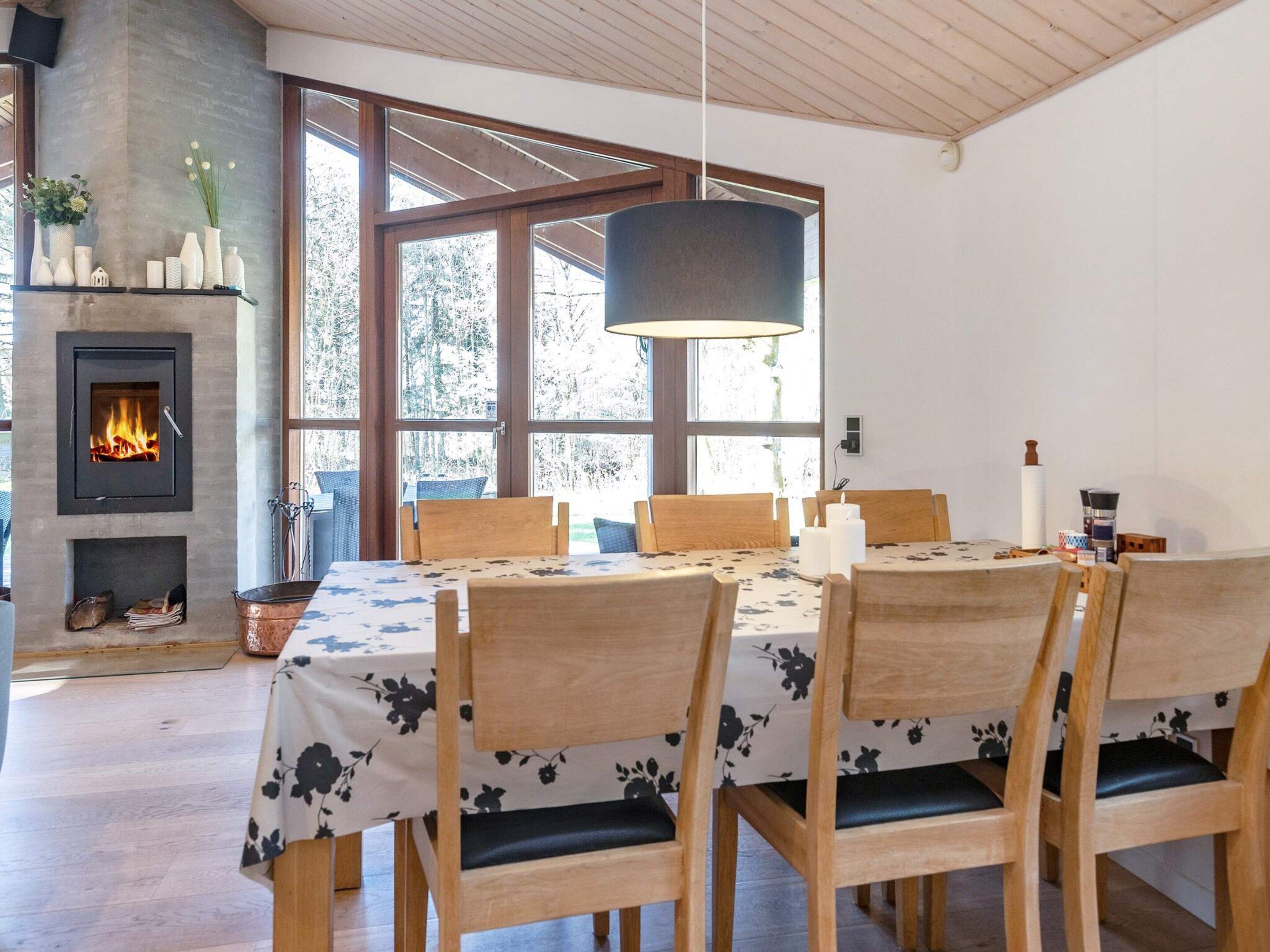 Un coin repas agrémenté d’une cheminée, de chaises en bois, d’une table recouverte d’une nappe à motifs floraux et de grandes fenêtres.