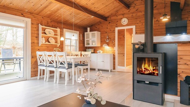 A cozy cabin interior with a wooden interior, a dining area with white chairs, a kitchen with white cabinets, and a wood-burning stove. - 4 Person Holiday Home in Jerup (Jerup)