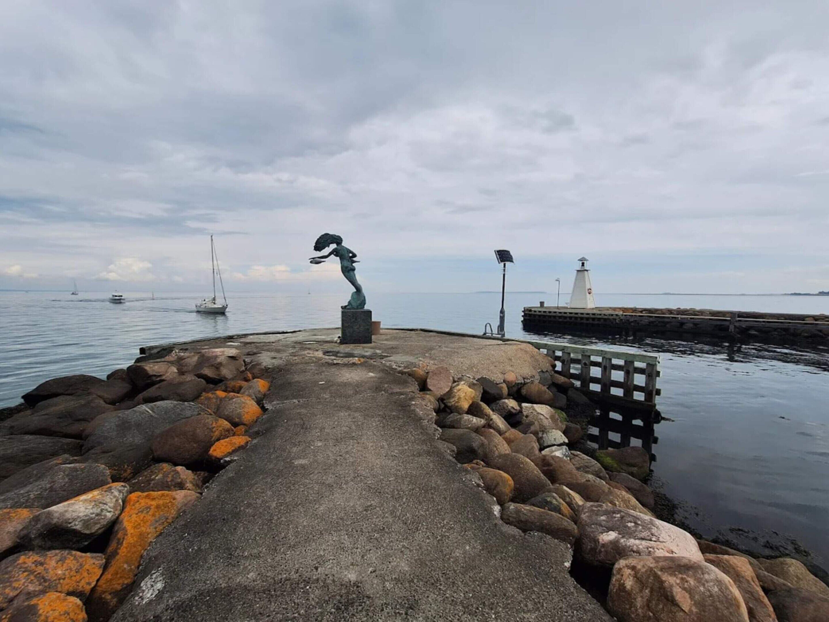Eine Küstenpromenade mit einer Statue, einem Leuchtturm und Booten in der Ferne.