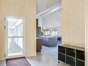 A modern kitchen with a central island, a dining area with chairs, and a hallway leading to a door with a view of a street.