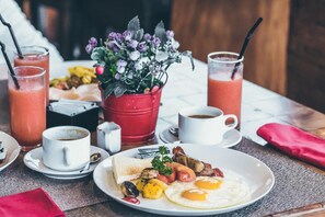 A breakfast setting with a plate of eggs, sausages, and vegetables, a red flower pot, and two glasses of a red beverage. - Deer Run Lodging House (Skaneateles)