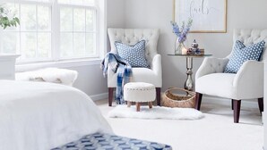 A bedroom with two white armchairs, a small table with a vase and candle, and a large window. - Deer Run Lodging House (Skaneateles)