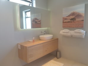 A modern bathroom with a wooden vanity, a white sink, a mirror, a small trash can, and a towel rack with folded towels. - Small Cottage With sep Bedroom, Specious Bathroom, Lounge and Open-plan Kitchen (Cape Town)