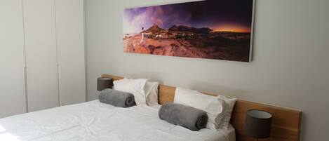 Un lit bien fait, avec du linge de lit blanc et deux serviettes grises enroulées, une tête de lit en bois et un tableau représentant un paysage montagneux accroché au mur.