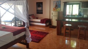 Ein Zimmer mit einem Bett, einer kleinen Küchenzeile, einer Essecke mit Hockern, einem roten Teppich und einem Deckenventilator.
