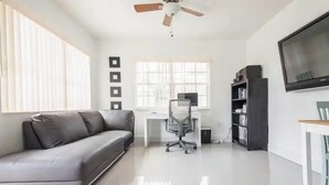 A modern living room with a grey sofa, a white desk, a black bookshelf, a TV, and a ceiling fan. - Miami Health District • 2 Gbps Internet • 75” & 55” TVs • Stainless Steel (Miami)