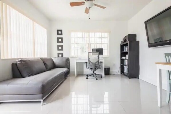 A modern living room with a grey sofa, a white desk, a black bookshelf, a TV, and a ceiling fan. - Miami Health District • 2 Gbps Internet • 75” & 55” TVs • Stainless Steel (Miami)