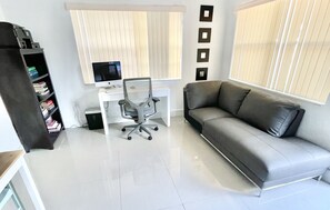A modern living room with a grey sofa, a white desk with a computer, and a bookshelf. - Miami Health District • 2 Gbps Internet • 75” & 55” TVs • Stainless Steel (Miami)
