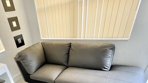 A modern, L-shaped gray sofa with a sleek design, positioned in a well-lit room with a large window and a white floor. - Miami Health District • 2 Gbps Internet • 75” & 55” TVs • Stainless Steel (Miami)