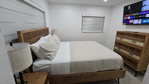 A bedroom with a bed, bedside lamp, wooden dresser, and a TV displaying various channels. - Miami Health District • 2 Gbps Internet • 75” & 55” TVs • Stainless Steel (Miami)